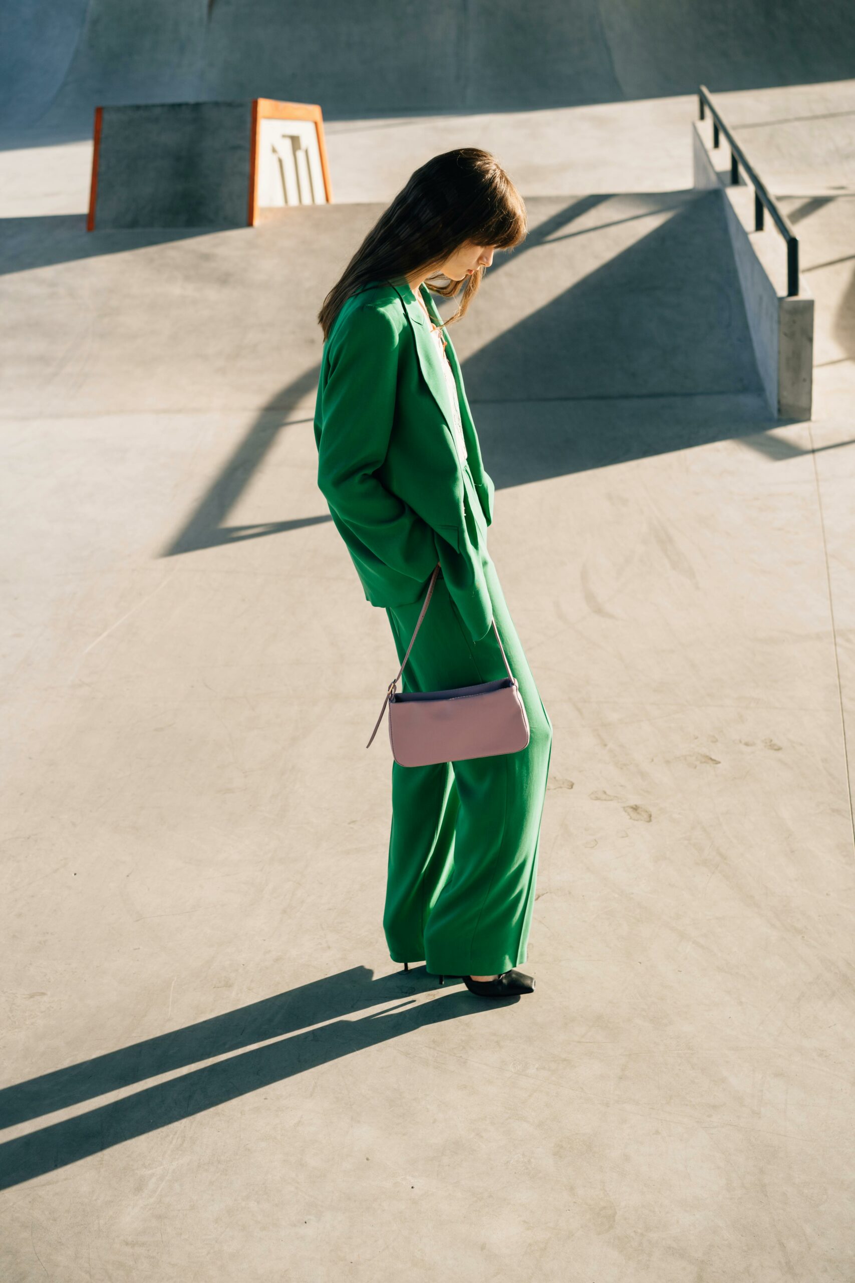 Elegant woman in a green suit with a pink bag striding in a modern outdoor setting.